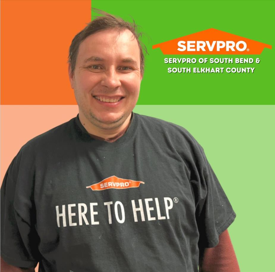 Man wearing a t shirt that says "Here to Help" smiles at camera with a multi colored background and a SERVPRO logo behind him